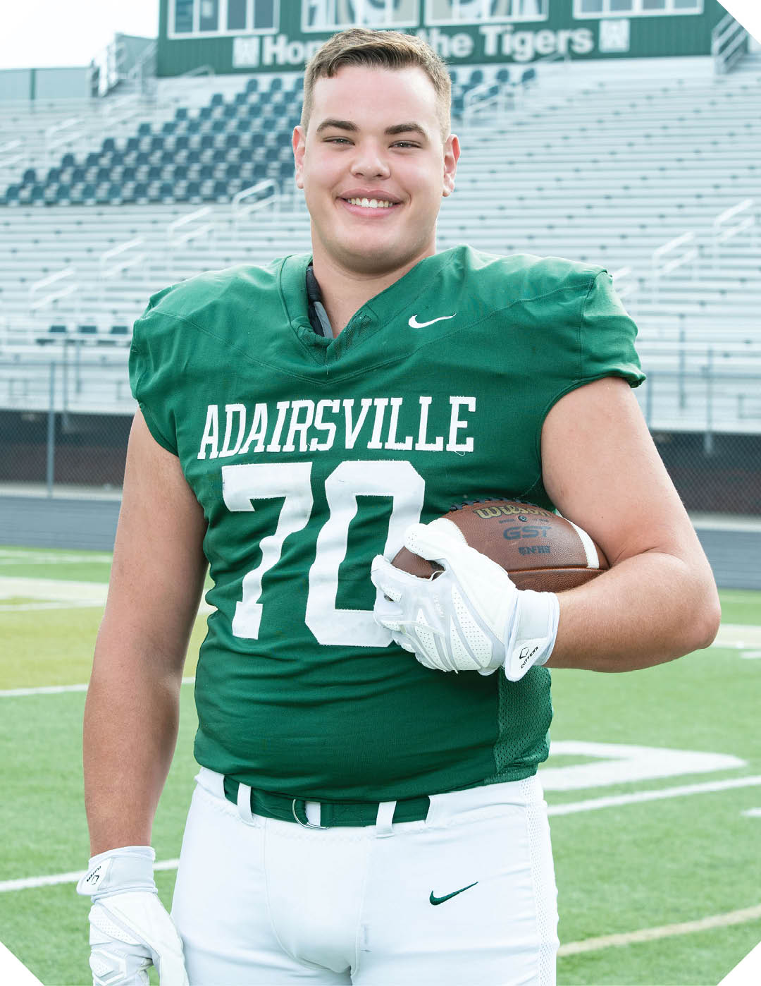 A person wearing a green football jersey with the number 70 and the word Adairsville is standing on a football field holding a football and smiling. Description generated by AI