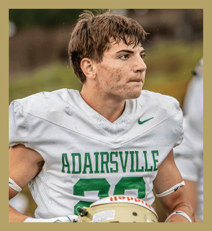A person wearing a white football jersey with “ADAIRSVILLE" and the number 22 in green is holding a gold football helmet decorated with paw prints and a large green "A". Description generated by AI