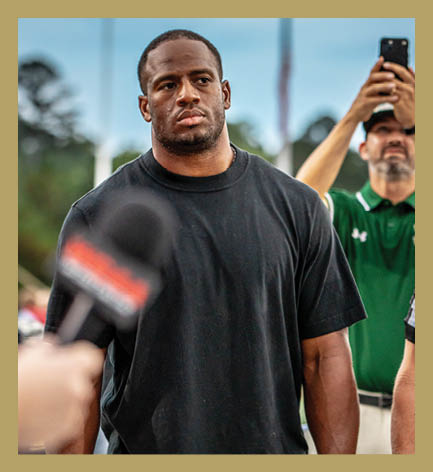 A person wearing a black t-shirt stands with a serious expression while another person in a green polo shirt holds up a phone in the background and a microphone is pointed toward the person in black. Description generated by AI