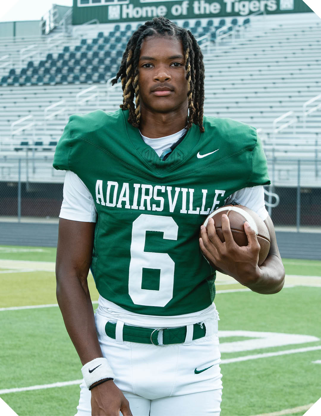 A person with shoulder-length dreadlocks wearing a green football jersey with the number 6 and the word Adairsville is standing on a football field holding a football. Description generated by AI