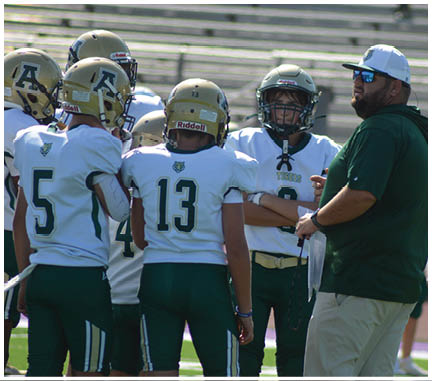 A group of people wearing football uniforms and one person in a green shirt and beige pants are standing together on a field. Description generated by AI