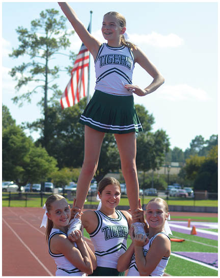 A person wearing a cheerleading outfit with the word “TIGERS" is smiling and raising one arm outdoors with an American flag in the background. Description generated by AI