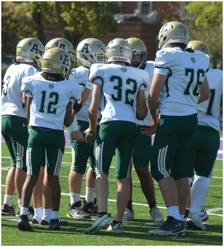 A group of people wearing football helmets and uniforms are gathered on a field, seemingly discussing strategy or preparing for a play. Description generated by AI