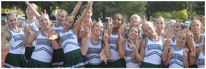 A group of people wearing matching cheerleading uniforms with green skirts and white tops stand closely together on a running track. Description generated by AI