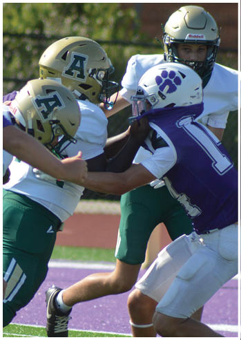Four people in football uniforms are engaged in a play on a green field with a purple track in the background. Description generated by AI