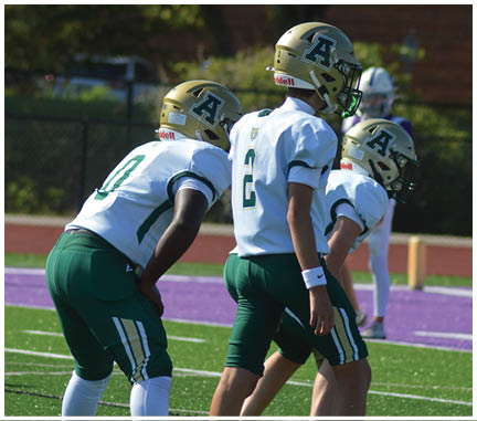 Three people wearing green and white sports uniforms stand on a football field with artificial turf and a purple track in the background. Description generated by AI