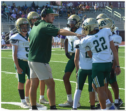 A group of people wearing football uniforms and helmets are gathered on a green field with white yard lines, with one person in shorts and a green shirt standing among them. Description generated by AI
