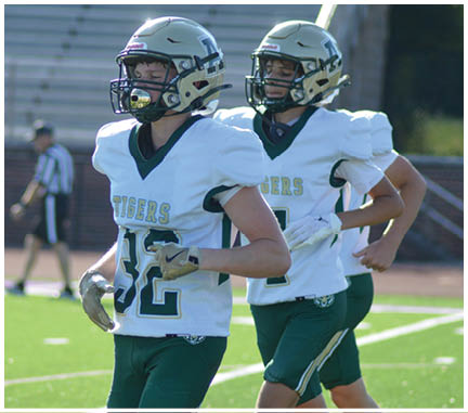Two people wearing green and white football uniforms are running on a grassy field. Description generated by AI