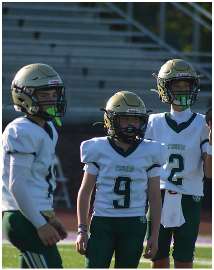 Three people wearing football helmets and white jerseys with green pants stand on a field, with one holding a football. Description generated by AI