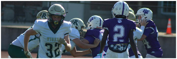 People wearing football uniforms are engaged in a game or practice on a field with one person in a white jersey running and others in purple and white jerseys nearby. Description generated by AI