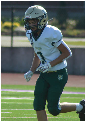 A person wearing a white football helmet and jersey with the number three is running on a green football field near a track and a fence. Description generated by AI