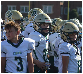 Children wearing football uniforms and helmets stand together on a field with a building in the background. Description generated by AI