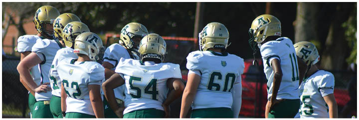 A group of people wearing white and green football uniforms and gold helmets are gathered closely together on a field. Description generated by AI