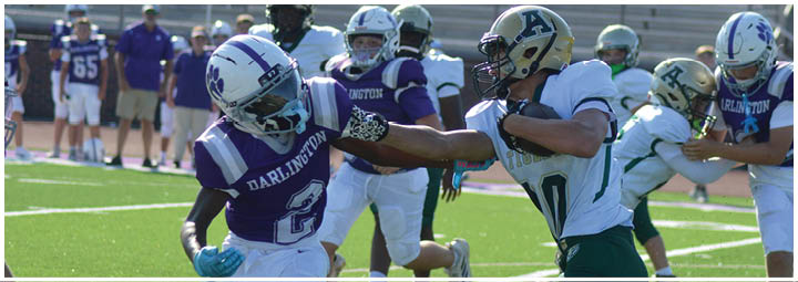People in football uniforms are actively engaged in a game on a sunny field with one player in white and green running while others in purple and white attempt to block or tackle. Description generated by AI