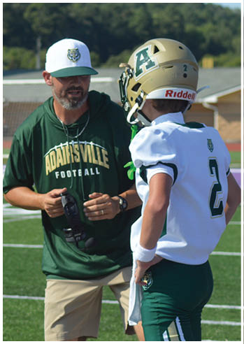 An adult wearing a green hoodie and cap is talking to a person in a white football uniform and helmet on a sports field. Description generated by AI