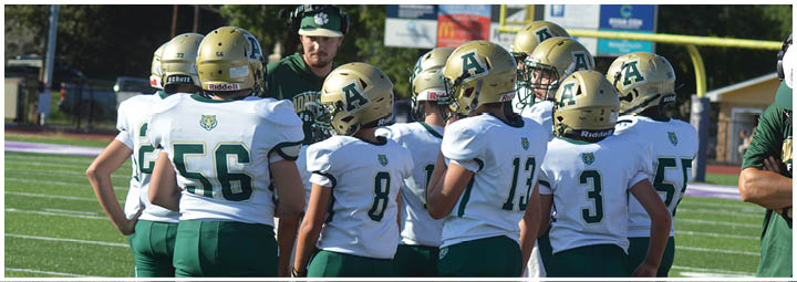 A group of people wearing white and green football uniforms with gold helmets are gathered on a sports field. Description generated by AI