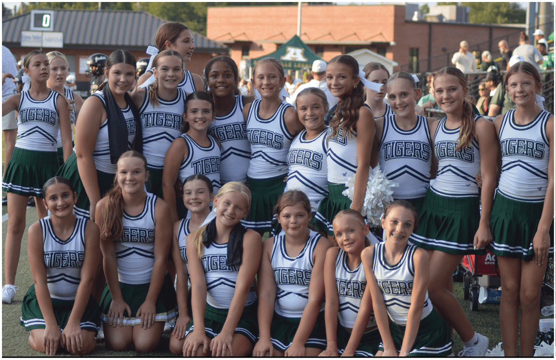 A group of smiling cheerleaders wearing matching green and white uniforms with “Tigers" on the front pose together on a sports field during daylight. Description generated by AI