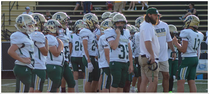 A group of people wearing football helmets and uniforms stand together on a field with a coach in a white shirt and green cap. Description generated by AI