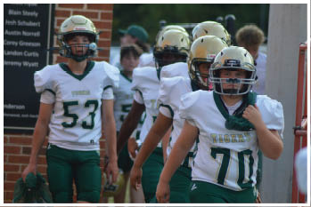 Two people wearing white football uniforms with green and gold accents and helmets stand on a field with a blurred background of other players and a sign. Description generated by AI