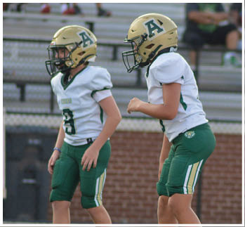 Two people wearing football uniforms and helmets stand on a field with one person in a ready stance and the other standing upright. Description generated by AI