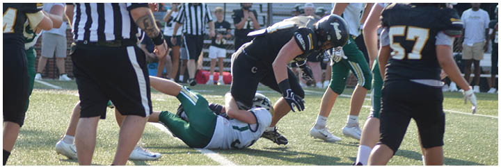 A group of people in sports uniforms are playing football on a field with one person tackling another while referees and other players observe. Description generated by AI