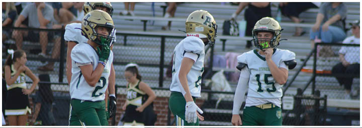 Three people in football uniforms and helmets stand on a field with cheerleaders and spectators in the background. Description generated by AI