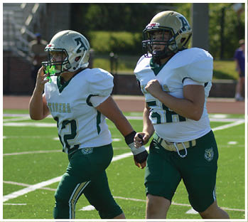 Two groups of people in football uniforms are engaged in a play on a green field with cheerleaders and a brick wall in the background. Description generated by AI