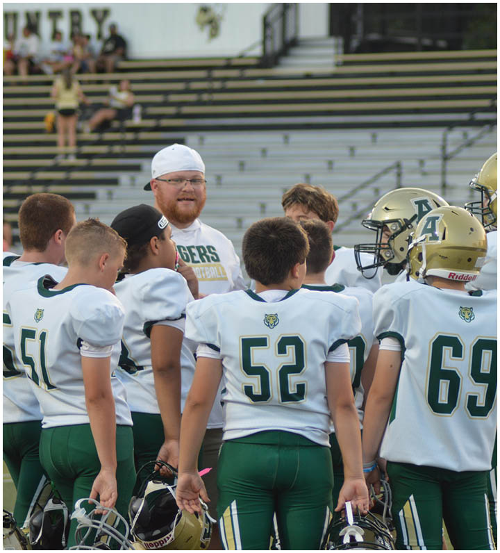 A group of people wearing football uniforms stand together on a field with bleachers and a sign that reads “COUNTRY" in the background. Description generated by AI