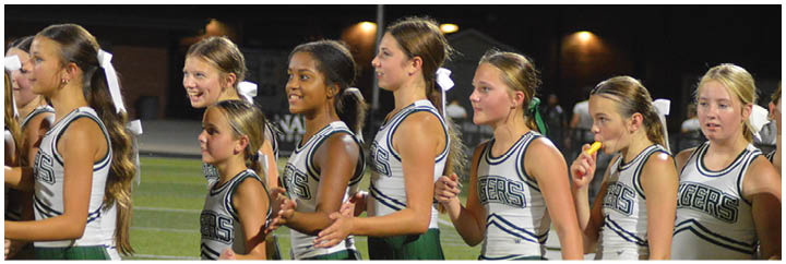 A group of people in cheerleading uniforms stand in a line on a sports field at night, some smiling and one eating a snack. Description generated by AI