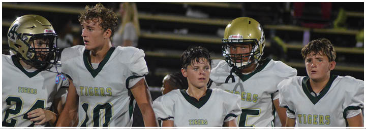 Four young people wearing white football uniforms with “Tigers" on the front stand closely together on a field, appearing focused and attentive. Description generated by AI