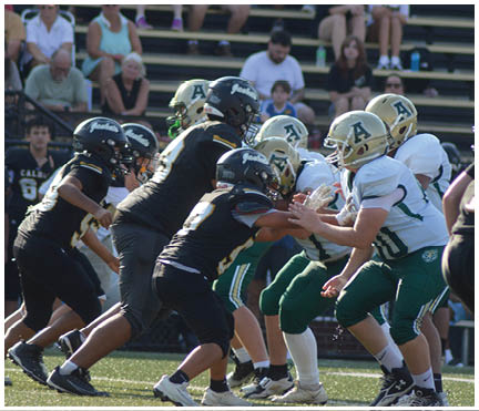 A group of people in football uniforms are engaged in a play on a field with spectators seated in bleachers behind them. Description generated by AI