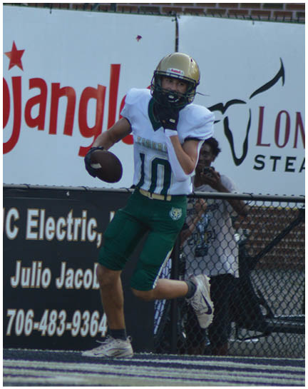 A person wearing a football uniform with a gold helmet and green pants is running on a field while holding a football near advertising signs. Description generated by AI