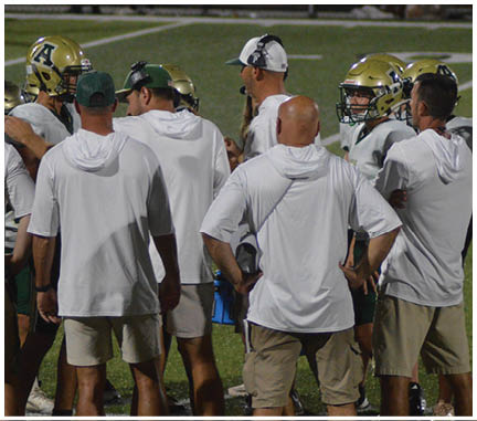 A group of people wearing white shirts and headsets are standing on a sports field, appearing to coach or direct players in football helmets. Description generated by AI
