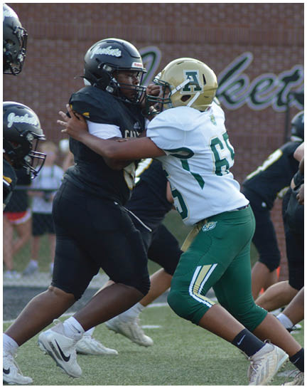 Two football players in helmets and uniforms are engaged in a blocking stance on a field during a game. Description generated by AI