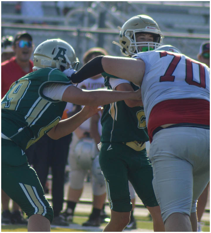 Three football players in green uniforms and helmets are engaged in a play against a player in a white and red uniform on a sunny field with bleachers and spectators in the background. Description generated by AI