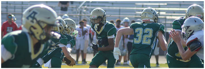People wearing football helmets and uniforms are engaged in a game or practice on a field with empty bleachers in the background. Description generated by AI
