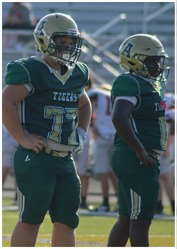 A person wearing a green football uniform with the number 77 and the word Tigers stands on a field with hands on hips. Description generated by AI