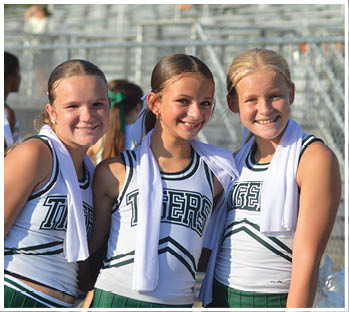 Three smiling people wearing matching cheerleading uniforms with towels draped around their necks stand close together outdoors in front of bleachers. Description generated by AI