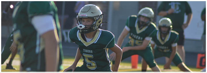 Children wearing football helmets and uniforms are on a field, with one child in the foreground looking focused and others in the background. Description generated by AI