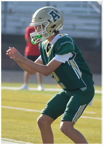 A person in a green football uniform and helmet is crouched in a ready position on a football field with another similarly dressed person partially visible nearby. Description generated by AI