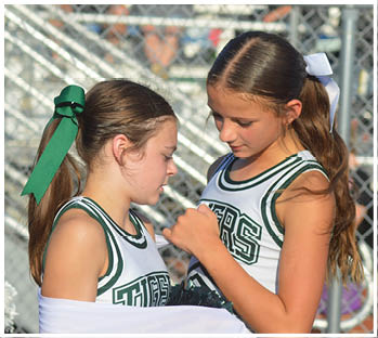 Two children in cheerleading uniforms are closely interacting, with one appearing to adjust or sign something on the other's outfit near a chain-link fence. Description generated by AI