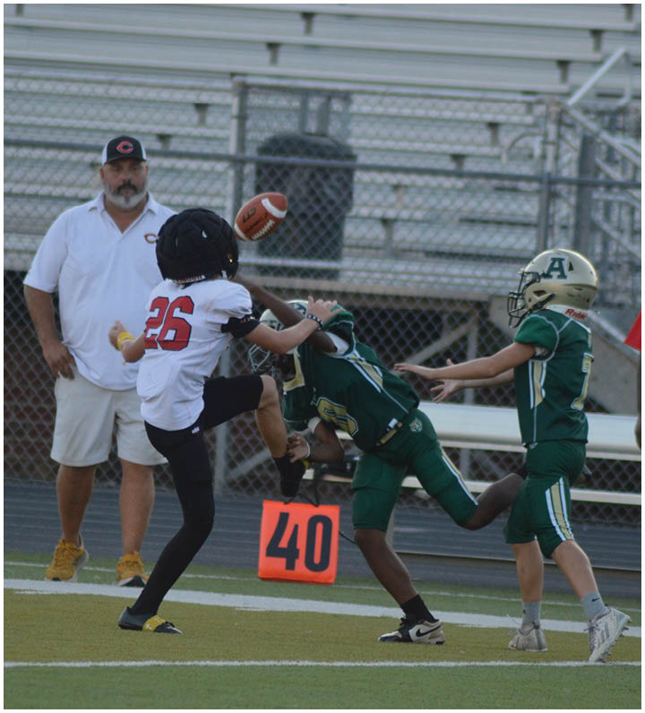 A person in a white sports uniform is kicking a football while two people in green football uniforms and helmets attempt to block the kick on a field with a coach standing in the background. Description generated by AI