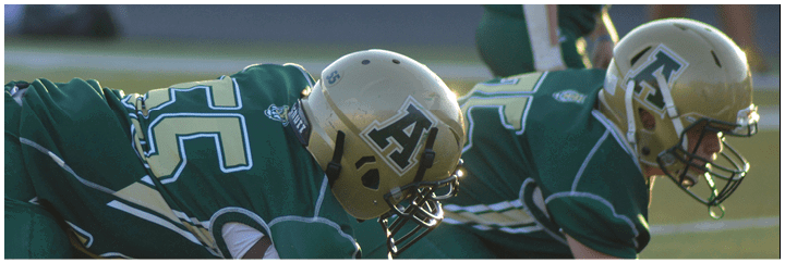 Three people in football uniforms and helmets are crouched in a ready position on a grassy field during a game or practice. Description generated by AI