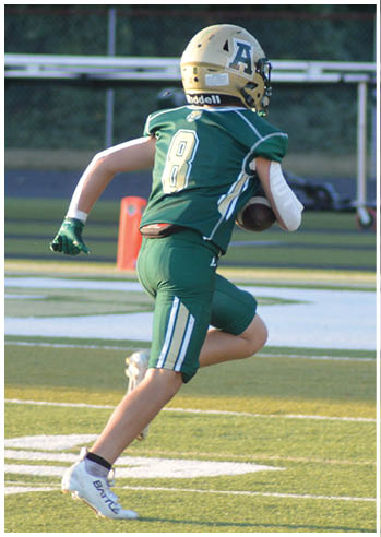 A person in green football gear and a gold helmet is running on a football field while holding a football. Description generated by AI