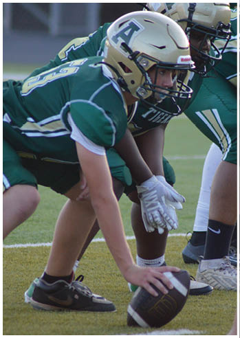 People in green football uniforms and helmets are lined up on a field with one person holding a football ready to snap it. Description generated by AI
