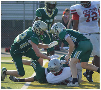 Several people in green football uniforms are surrounding and tackling a person in a white football uniform on a sunlit field during a game. Description generated by AI