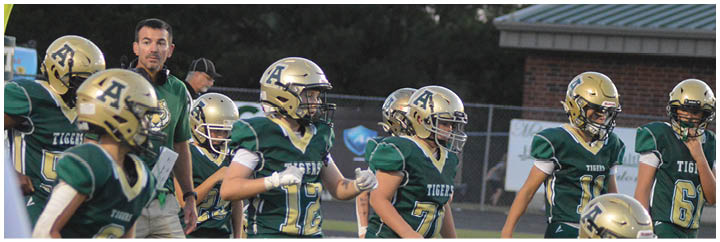 A group of people wearing green and gold football uniforms and helmets are standing together on a field with a coach holding a clipboard nearby. Description generated by AI