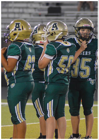 Two people in green and gold football uniforms and helmets stand on a field with empty bleachers in the background. Description generated by AI