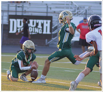 Two people in green football uniforms and gold helmets are on a field with one holding a football in place while the other prepares to kick it. Description generated by AI