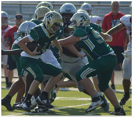 Several people wearing football uniforms are actively engaged in a play on a sunlit field with one person holding the ball and others blocking or attempting to tackle. Description generated by AI